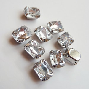 May include: A group of eight clear, faceted, rectangular rhinestones with silver settings. The rhinestones are arranged in a loose cluster on a white background.
