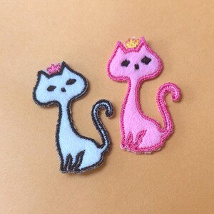 K&ouml;nnte beinhalten: Zwei Katzen-Applikationen aus Filz. Eine ist hellblau mit schwarzer Umrandung und einer rosa Krone. Die andere ist rosa mit rosa Umrandung und einer goldenen Krone. Beide haben schwarze Augen und sitzen auf hellorangefarbenem Hintergrund.