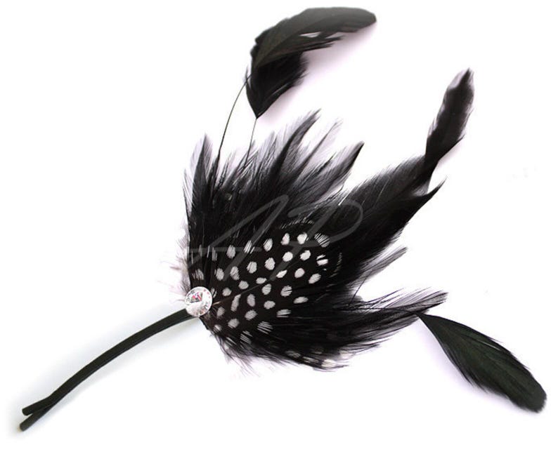 May include: A black feather hair accessory with a central cluster of feathers featuring white polka dots. A small, clear jewel is set in the center, and the accessory has a thin, black stem.