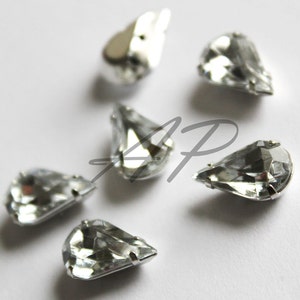 May include: Six clear teardrop-shaped crystal rhinestones with silver settings. The rhinestones are arranged in a semi-circle on a white background.