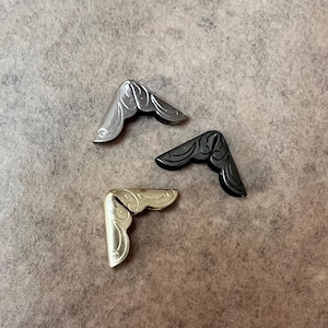 May include: Three metal corner protectors with a decorative design. The corner protectors are in silver, gold, and black.