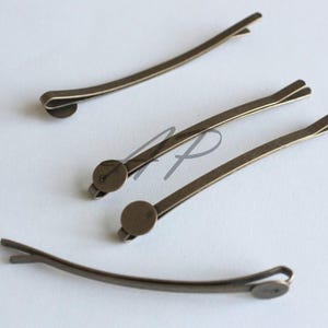 May include: Four antique bronze-colored bobby pins. Each pin has a curved, slender design with a small, round decorative element at one end. The pins are arranged on a light-colored surface, showcasing their simple elegance.