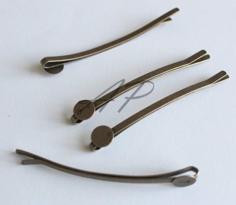 May include: Four antique bronze-colored bobby pins. Each pin has a curved, slender design with a small, round decorative element at one end. The pins are arranged on a light-colored surface, showcasing their simple elegance.