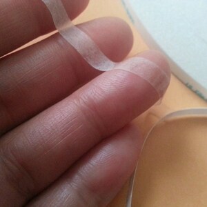 Pode incluir: Um rolo de fita adesiva dupla face transparente, com uma extremidade da fita sendo segurada por um dedo.