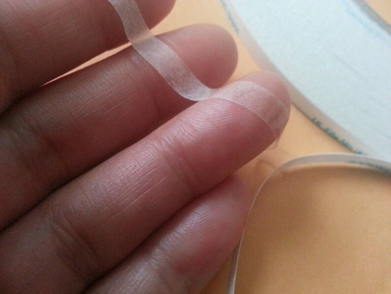 Pode incluir: Um rolo de fita adesiva dupla face transparente, com uma extremidade da fita sendo segurada por um dedo.