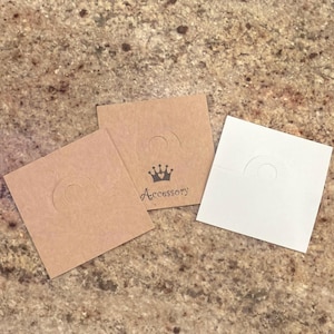 May include: Three square earring display cards. Two are brown, one is white. Each card has a circular cutout and a curved top. One brown card has a crown and the word "Accessory" printed in black.