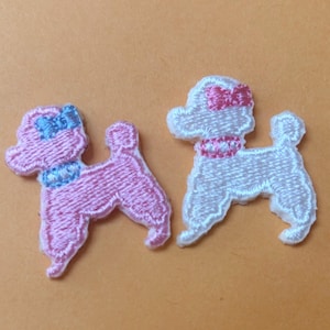 May include: Two embroidered poodle patches. One is pink with a blue bow and collar, the other is white with a pink bow and collar. The patches are detailed with stitched outlines and are likely for clothing or craft projects.