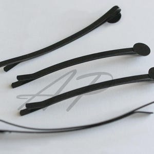 May include: Four black bobby pins arranged on a white surface. Three pins have a rounded end, and one is open. The pins are slightly curved and appear to be made of metal. These hair accessories are designed to secure hair.