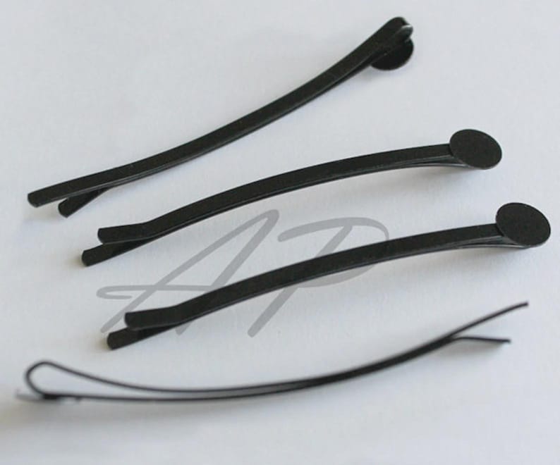 May include: Four black bobby pins arranged on a white surface. Three pins have a rounded end, and one is open. The pins are slightly curved and appear to be made of metal. These hair accessories are designed to secure hair.