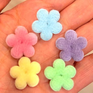 May include: Five small, plush flower embellishments in various pastel colors: pink, light blue, purple, yellow, and green. Each flower has five rounded petals and a soft, fuzzy texture. These are likely craft supplies.