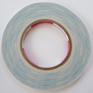 Pode incluir: Um rolo de fita adesiva de dupla face azul e branca com um r&oacute;tulo rosa. A fita est&aacute; sobre um fundo branco.