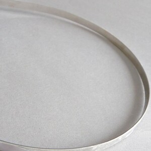 May include: A thin, flexible, silver-colored metal strip curved into a partial oval shape. The material has a smooth, reflective surface with a consistent width throughout its length, forming a simple, open-ended loop structure.