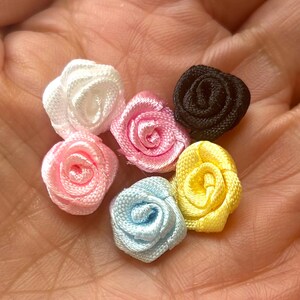 May include: A collection of small, fabric rose embellishments in various colors, including white, pink, black, yellow, and blue. These miniature roses are tightly wound and appear to be made of satin or a similar material, perfect for craft projects.