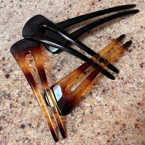 May include: Two hair forks in the image. One is black, and the other is a tortoiseshell pattern of brown and amber. The forks have a curved design and are made of a glossy material. They are likely used for styling hair.