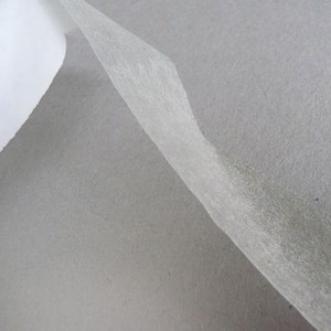 May include: A thin, translucent, off-white adhesive tape strip is partially peeled away from a solid, matte gray paper backing. The tape has a fibrous, semi-transparent texture, while the underlying paper surface appears smooth and uniform.