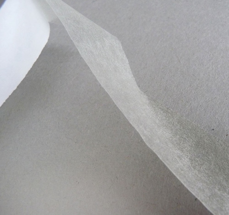 May include: A thin, translucent, off-white adhesive tape strip is partially peeled away from a solid, matte gray paper backing. The tape has a fibrous, semi-transparent texture, while the underlying paper surface appears smooth and uniform.