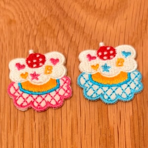 May include: Two embroidered cupcake patches. One is pink and the other is blue. Each patch has a white frosting top with a red strawberry, pink hearts, and a gold letter "B". The bottom of each patch has a scalloped edge.