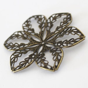 May include: A bronze-colored metal flower charm with a delicate, intricate design. The charm is made of thin metal and has a filigree pattern.