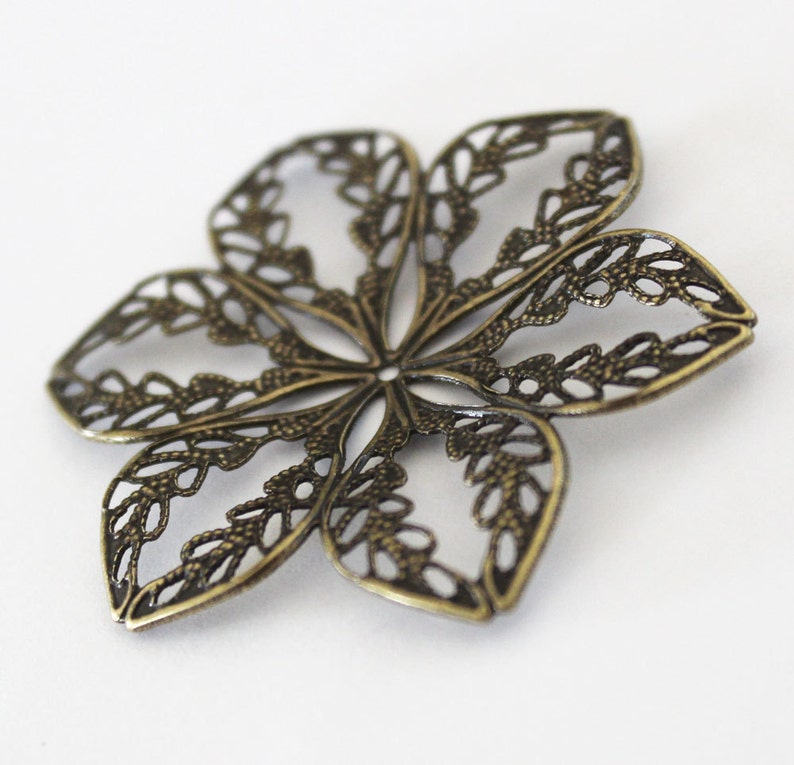 May include: A bronze-colored metal flower charm with a delicate, intricate design. The charm is made of thin metal and has a filigree pattern.