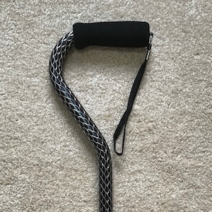 May include: A black and silver walking cane with a padded handle and a wrist strap. The cane has a decorative pattern of scales.