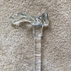 May include: A clear, decorative walking cane with a curved handle. The handle and shaft are made of a transparent material, giving it a sleek and modern appearance. The cane's design is both functional and stylish, suitable for those seeking mobility assistance or a fashion accessory.
