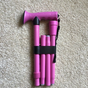 Könnte beinhalten: Ein rosa zusammenklappbarer Gehstock mit schwarzem Griff und schwarzem Band. Der Stock ist zusammengeklappt und mit einem schwarzen Band befestigt.