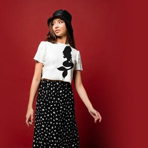 Puede incluir: Una mujer con un top blanco corto con un dise&ntilde;o de silueta de sirena negra, una falda de lunares negros y blancos y un sombrero de cubo negro. Est&aacute; de pie frente a un fondo rojo.