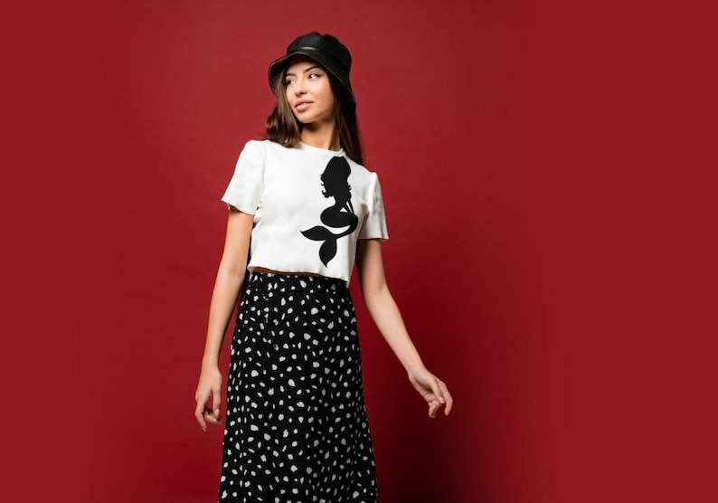 Puede incluir: Una mujer con un top blanco corto con un dise&ntilde;o de silueta de sirena negra, una falda de lunares negros y blancos y un sombrero de cubo negro. Est&aacute; de pie frente a un fondo rojo.