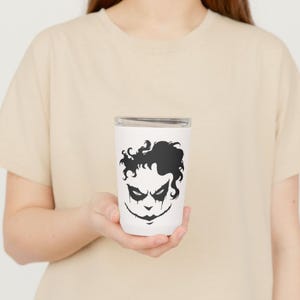 May include: White tumbler featuring a black graphic of a stylized face with dark eye makeup and a sinister smile. The tumbler has a silver lid and is held by a person wearing a beige t-shirt. The design is reminiscent of a comic book villain.