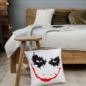 May include: White square pillow with a black and red graphic of a smiling face. The pillow is on a wooden floor in front of a bed with a wooden frame. A small wooden stool is to the left of the pillow.