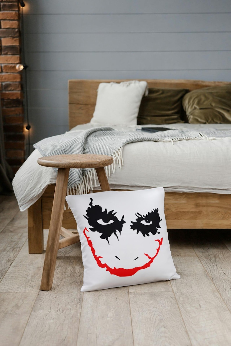 May include: White square pillow with a black and red graphic of a smiling face. The pillow is on a wooden floor in front of a bed with a wooden frame. A small wooden stool is to the left of the pillow.