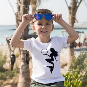 Puede incluir: Un ni&ntilde;o peque&ntilde;o con gafas de sol azules y una camiseta blanca con un gr&aacute;fico de sirena negra. La camiseta tiene un c&iacute;rculo blanco alrededor de la cabeza de la sirena.