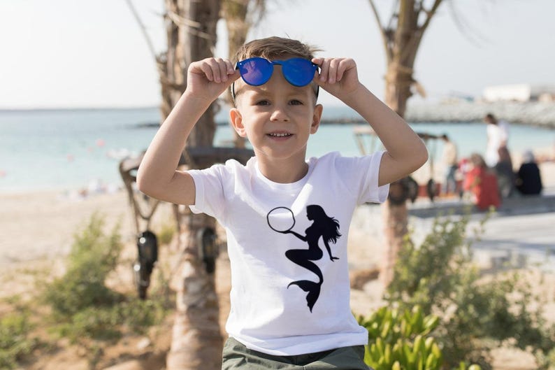 Puede incluir: Un ni&ntilde;o peque&ntilde;o con gafas de sol azules y una camiseta blanca con un gr&aacute;fico de sirena negra. La camiseta tiene un c&iacute;rculo blanco alrededor de la cabeza de la sirena.