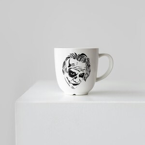 May include: White ceramic mug featuring a black and white illustration of a character's face. The mug has a curved handle and a rounded body. The design is a stylized portrait. The mug is sitting on a white surface.