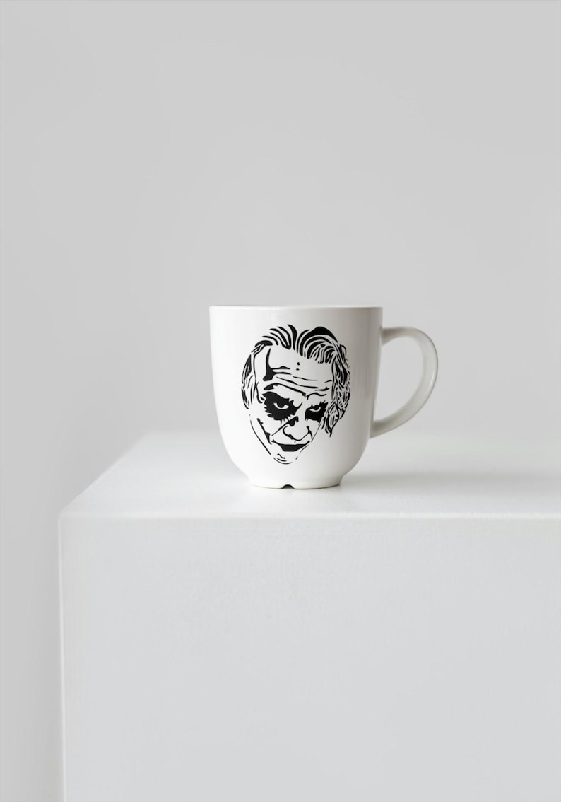 May include: White ceramic mug featuring a black and white illustration of a character's face. The mug has a curved handle and a rounded body. The design is a stylized portrait. The mug is sitting on a white surface.