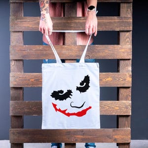May include: A white tote bag featuring a graphic of a smiling face with black eye shapes and a red smile. The bag has white straps and is held in front of a wooden pallet.