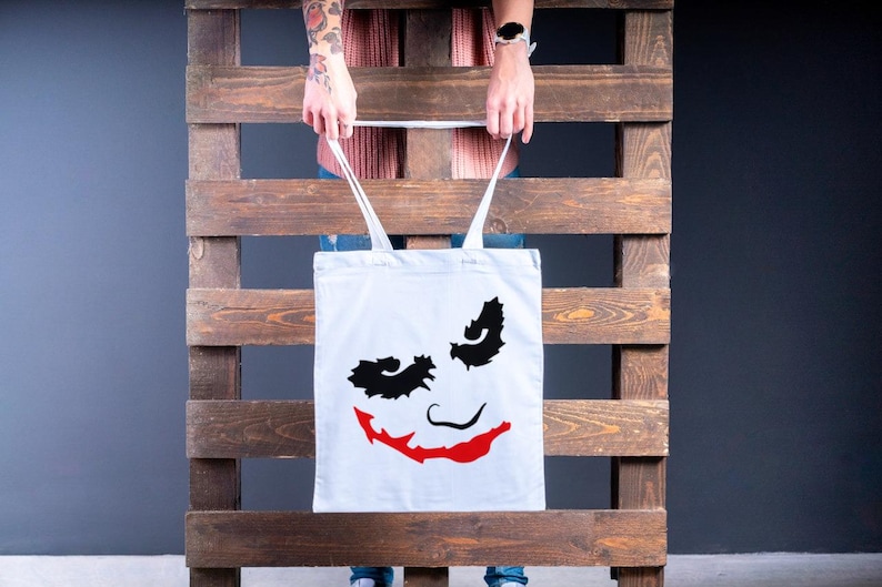May include: A white tote bag featuring a graphic of a smiling face with black eye shapes and a red smile. The bag has white straps and is held in front of a wooden pallet.