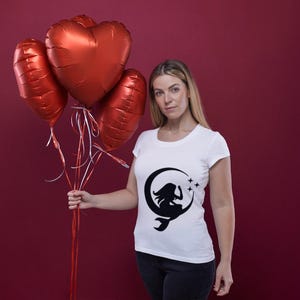 Puede incluir: Una mujer lleva una camiseta blanca con una silueta negra de una mujer sentada en una luna creciente con estrellas. Est&aacute; sosteniendo un ramo de globos rojos en forma de coraz&oacute;n.