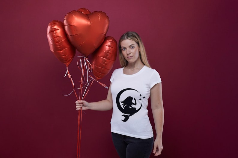 Puede incluir: Una mujer lleva una camiseta blanca con una silueta negra de una mujer sentada en una luna creciente con estrellas. Est&aacute; sosteniendo un ramo de globos rojos en forma de coraz&oacute;n.