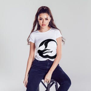 Puede incluir: Una camiseta blanca con un gr&aacute;fico de silueta de sirena negra en la parte delantera. La modelo lleva pantalones vaqueros azules y est&aacute; sentada en un taburete.
