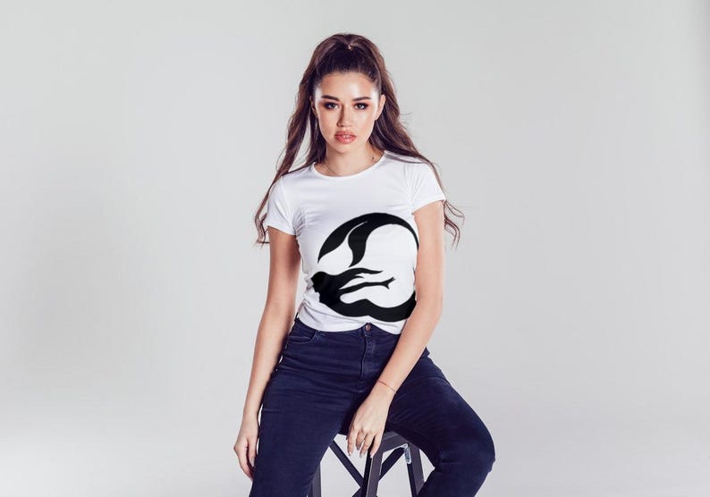 Puede incluir: Una camiseta blanca con un gr&aacute;fico de silueta de sirena negra en la parte delantera. La modelo lleva pantalones vaqueros azules y est&aacute; sentada en un taburete.