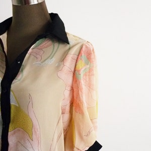 Puede incluir: Una blusa beige con cuello negro y estampado floral. Las mangas son abullonadas y tienen un ribete negro.