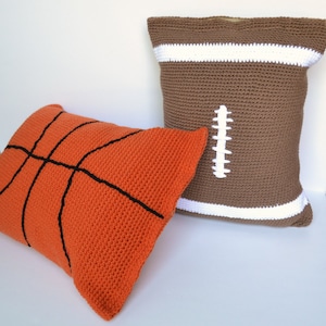 May include: Two crocheted pillows, one orange with a black basketball design and one brown with a white football design.
