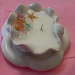 May include: A white seashell-shaped candle with a white wick, surrounded by seashells, starfish, and beads. The candle is sitting on a white seashell-shaped base.