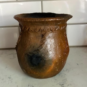 May include: A small, hand-formed brown clay pot with a wide, flared rim and a textured, uneven surface. The vessel features a dark, charred-looking circular patch on its side and decorative, raised clay markings along the upper rim and body.