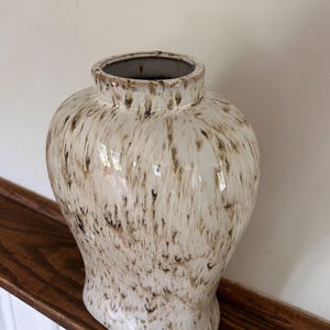 May include: A ceramic vase with a wide, rounded body and a short, narrow neck. The surface features a glossy white glaze with a mottled, streaky brown pattern that resembles dripping paint or natural stone textures throughout the entire exterior.