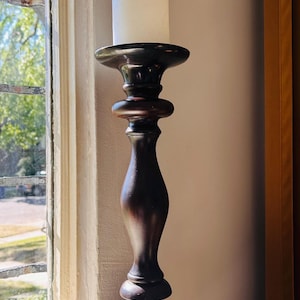 May include: A tall, dark brown wooden candlestick holder with a smooth, turned silhouette and a wide circular base. A plain, cylindrical white pillar candle is positioned securely in the top cup of the holder, creating a vertical display.