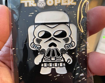May include: A white enamel pin featuring a cartoon skull trooper from the Star Wars universe. The pin has a black outline and blue details. The text "SKULL TROOPER" is written in gold letters above the trooper.