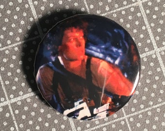 May include: A round button featuring a close-up image of a man from the movie "Aliens". The man is wearing a dark vest and has a red glow on his face. The button is white with a black border.