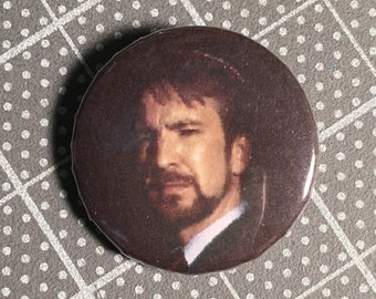 May include: A round button featuring a close-up portrait of a man with a beard and dark hair. The background is a dark brown color. The button is a promotional item for "The Button Depot".